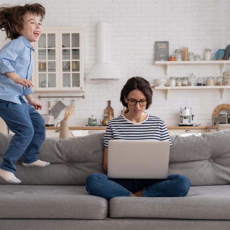 Eine weiße Frau sitzt im Schneidersitz und mit Laptop auf den Beinen auf einem grauen Sofa. Neben ihr springt ein kleines Kind auf dem Sofa auf und ab.