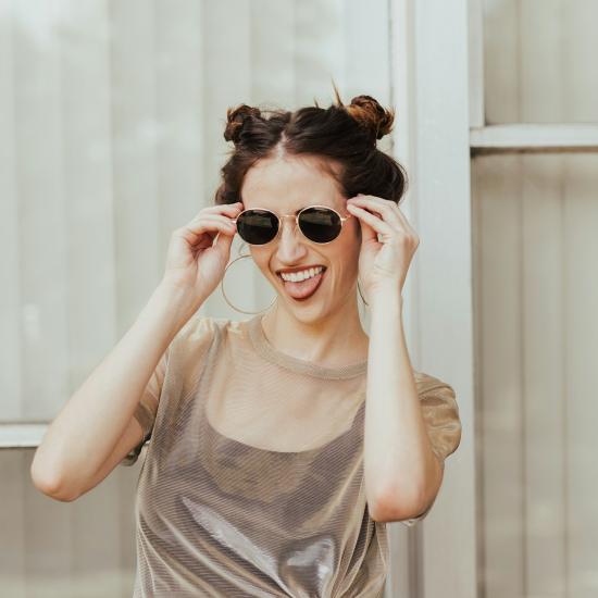 Eine junge Frau in einem dünnen Top und mit zwei Knubbelzöpfen greift lächelnd nach ihrer Sonnenbrille.